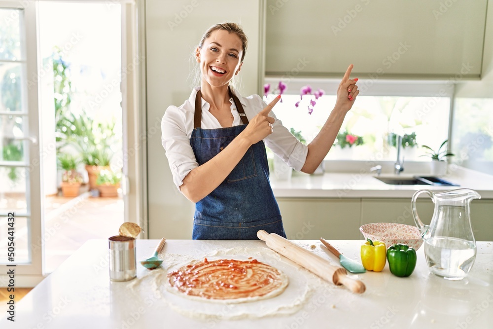 Beautiful blonde woman wearing apron cooking pizza smiling and looking at the camera pointing with two hands and fingers to the side.