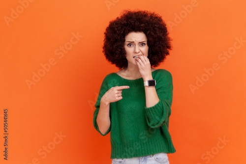 Woman with Afro hairstyle wearing green sweater showing watch on her hand, reminding of time, late hour, warning about deadline, biting fingers. Indoor studio shot isolated on orange background.