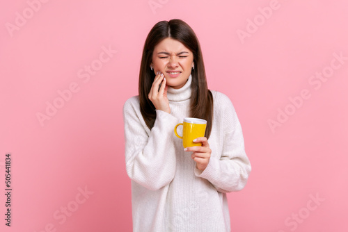 Sick brunette woman suffering from sensitive teeth after drinking hot or cold beverage, cavities, wearing white casual style sweater. Indoor studio shot isolated on pink background. photo