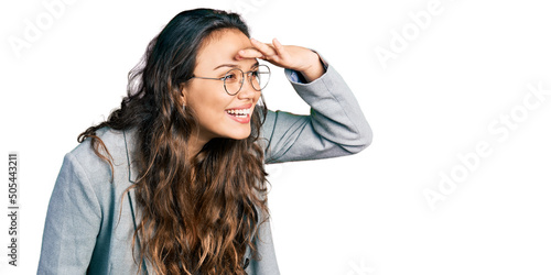 Young hispanic girl wearing business clothes and glasses very happy and smiling looking far away with hand over head. searching concept.