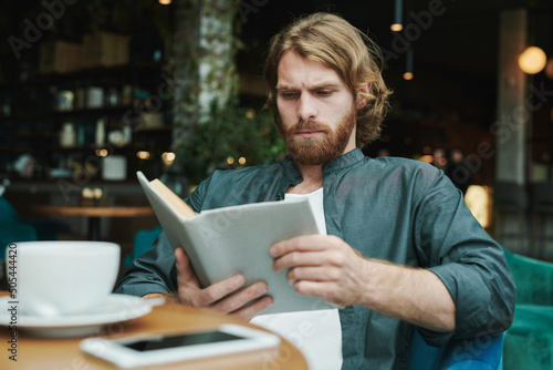 Serious handsome guy with beard sitting at table in cafe and drinking coffee while reading mail on smartphone
