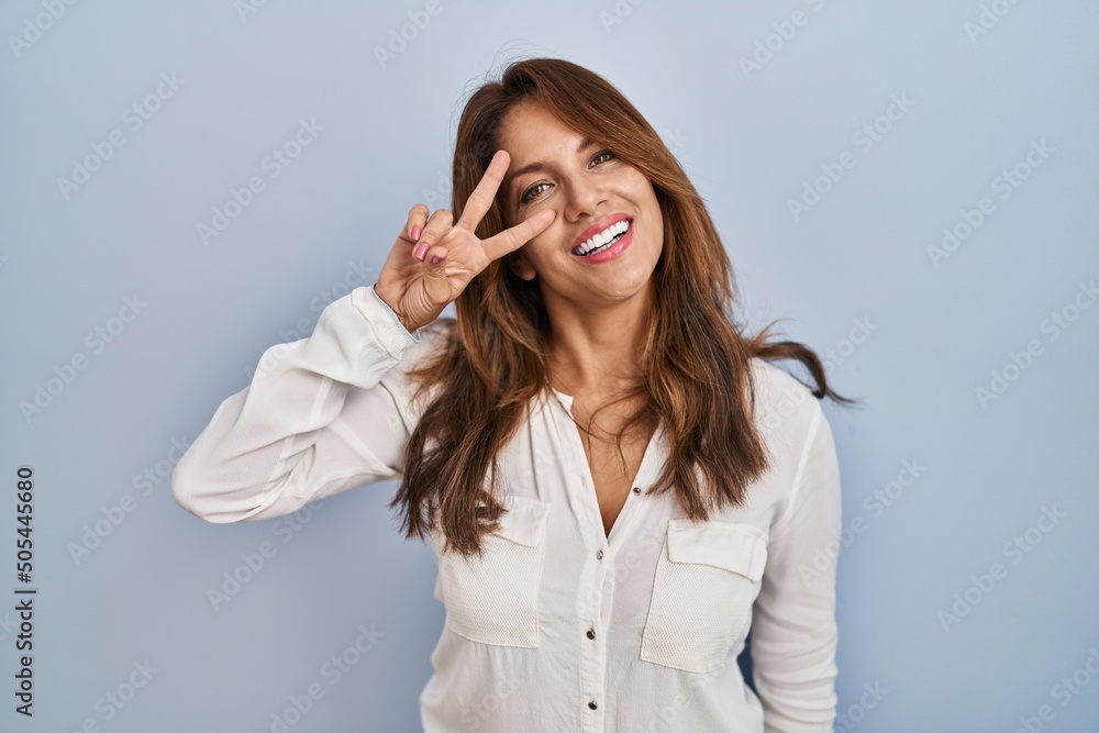 Hispanic woman standing over isolated background doing peace symbol with fingers over face, smiling cheerful showing victory Hispanic woman standing over isolated background doing peace symbol with fingers over face, smiling cheerful showing victory