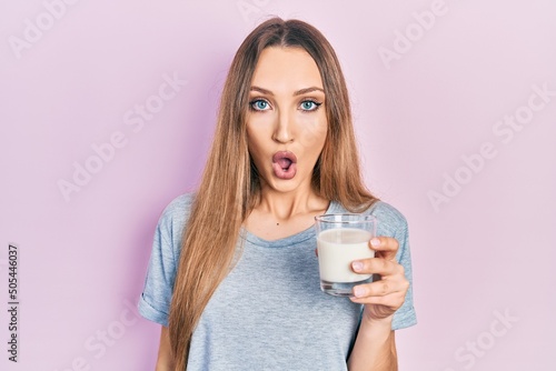 Young blonde girl holding glass of milk scared and amazed with open mouth for surprise, disbelief face