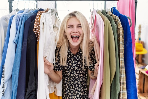Young blonde woman searching clothes on clothing rack celebrating crazy and amazed for success with open eyes screaming excited.