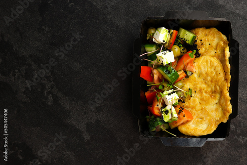 Healthy food delivery concept. Chicken schnitzel with Greek salad in an eco paper container on a black background. Restaurant dish delivery. Top view. Free space for text. photo