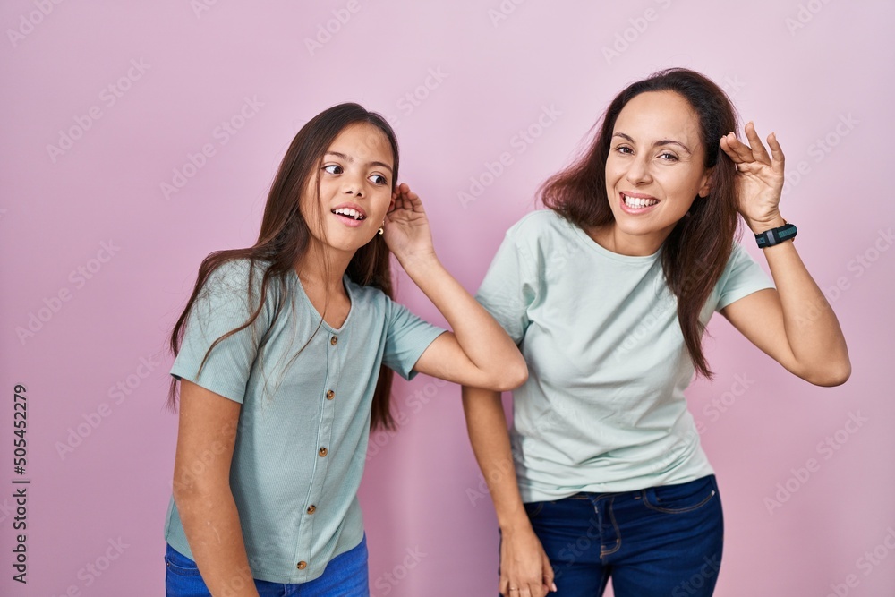 Young mother and daughter standing over pink background smiling with hand over ear listening an hearing to rumor or gossip. deafness concept. Young mother and daughter standing over pink background smiling with hand over ear listening an hearing to rumor or gossip. deafness concept.
