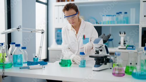 Middle age hispanic woman wearing scientist uniform pouring liquid on test tube at laboratory