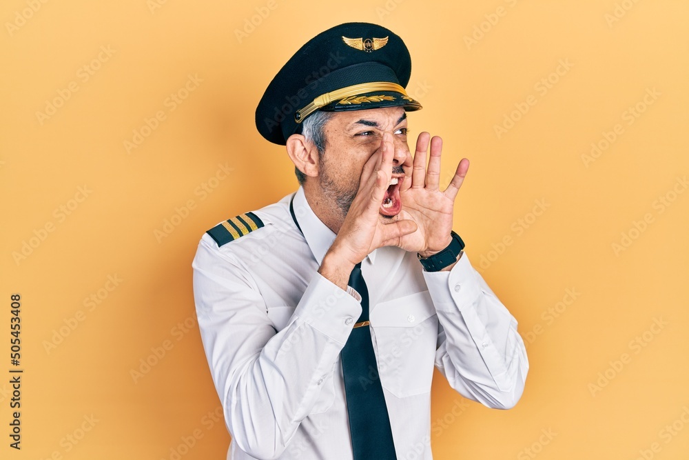 Handsome middle age man with grey hair wearing airplane pilot uniform shouting angry out loud with hands over mouth Handsome middle age man with grey hair wearing airplane pilot uniform shouting angry out loud with hands over mouth