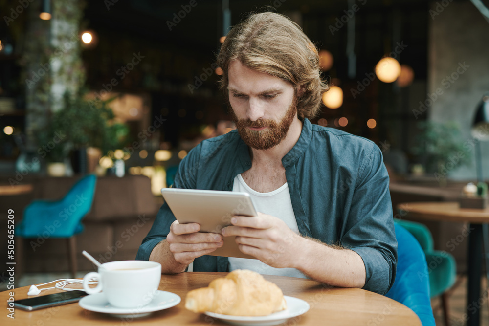 Young bearded manager sitting at table with coffee cup and croissant and recording voice message for colleague using smartphone in cafe Young bearded manager sitting at table with coffee cup and croissant and recording voice message for colleague using smartphone in cafe