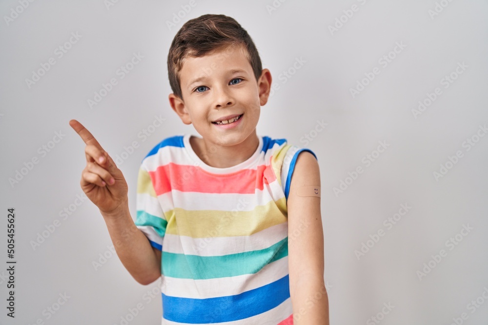 Young caucasian kid getting vaccine showing arm with band aid smiling happy pointing with hand and finger to the side Young caucasian kid getting vaccine showing arm with band aid smiling happy pointing with hand and finger to the side