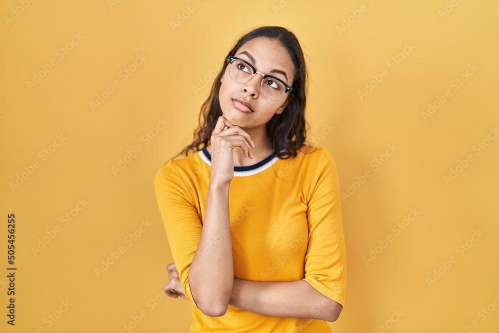Young brazilian woman wearing glasses over yellow background serious face thinking about question with hand on chin, thoughtful about confusing idea Young brazilian woman wearing glasses over yellow background serious face thinking about question with hand on chin, thoughtful about confusing idea