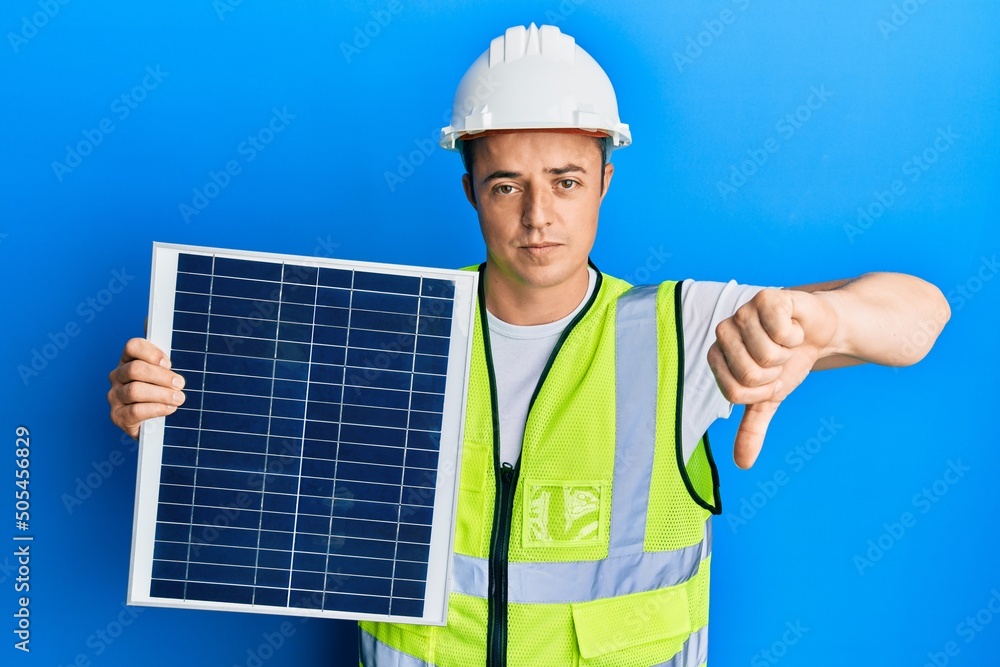 Handsome young man holding photovoltaic solar panel with angry face, negative sign showing dislike with thumbs down, rejection concept Handsome young man holding photovoltaic solar panel with angry face, negative sign showing dislike with thumbs down, rejection concept