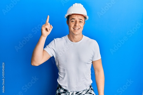 Handsome young man wearing builder uniform and hardhat pointing finger up with successful idea. exited and happy. number one.
