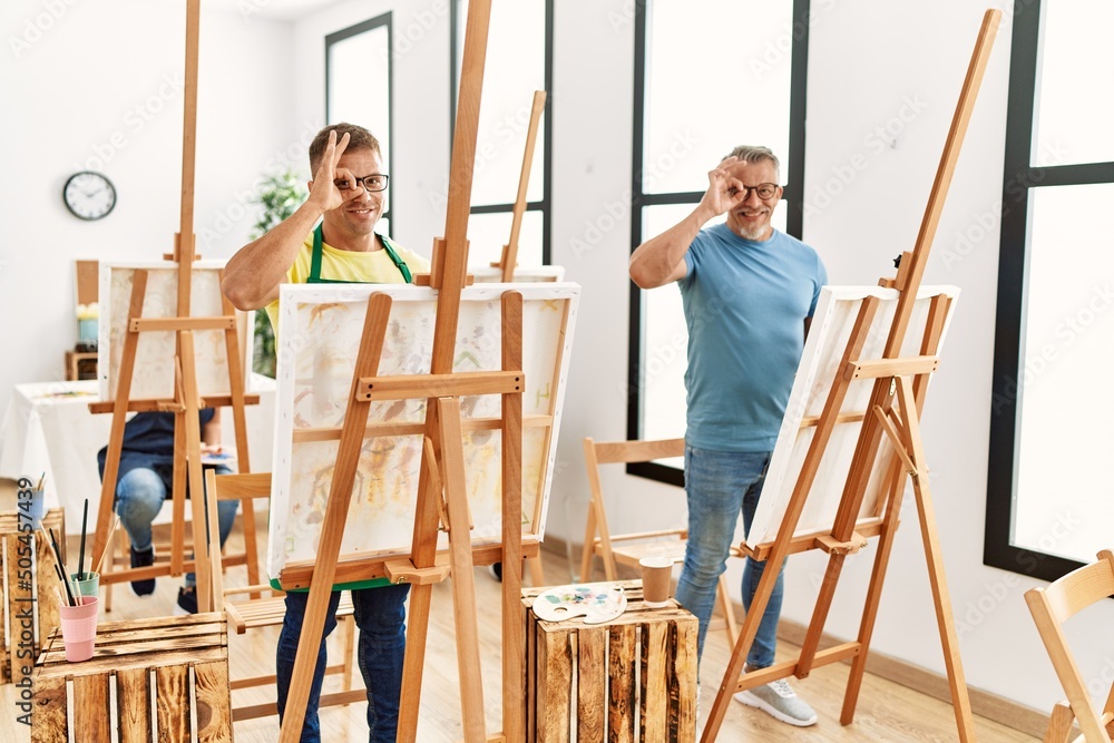 Group of middle age people artist at art studio smiling happy doing ok sign with hand on eye looking through fingers