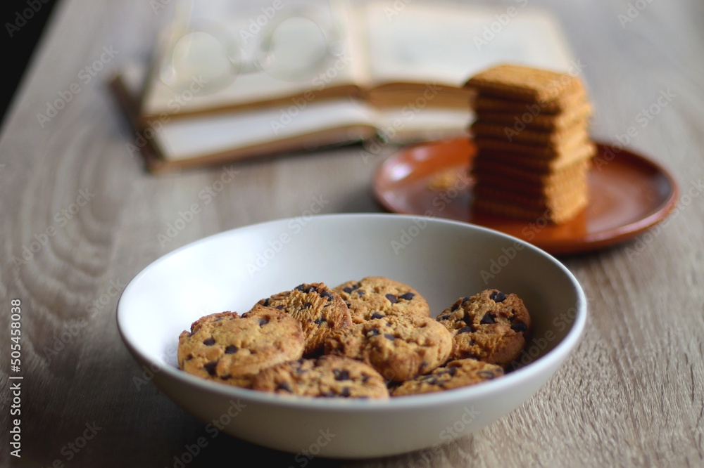 Bowl of chocolate chip cookies, plate of biscuits, open books and reading glasses on the table. Selective focus. Bowl of chocolate chip cookies, plate of biscuits, open books and reading glasses on the table. Selective focus.
