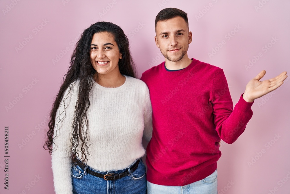 Young hispanic couple standing over pink background smiling cheerful presenting and pointing with palm of hand looking at the camera. Young hispanic couple standing over pink background smiling cheerful presenting and pointing with palm of hand looking at the camera.