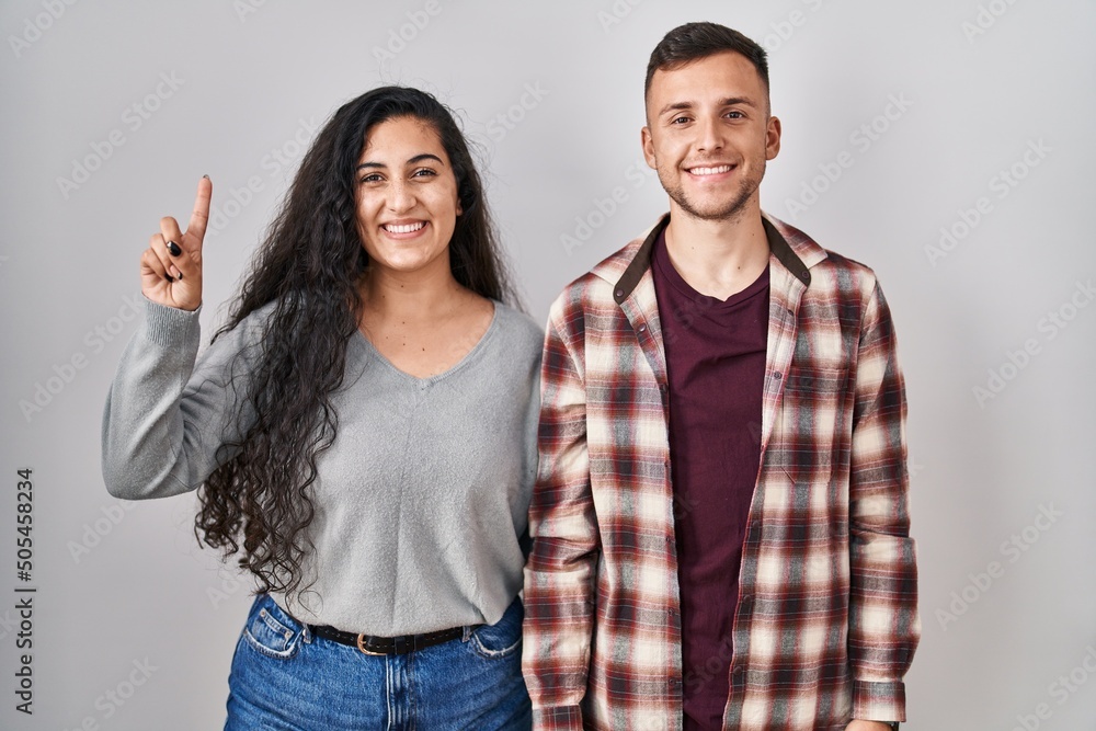 Young hispanic couple standing over white background showing and pointing up with finger number one while smiling confident and happy. Young hispanic couple standing over white background showing and pointing up with finger number one while smiling confident and happy.