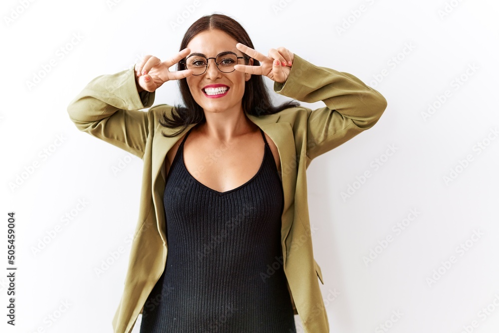Young brunette woman standing over isolated background doing peace symbol with fingers over face, smiling cheerful showing victory Young brunette woman standing over isolated background doing peace symbol with fingers over face, smiling cheerful showing victory