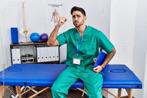 Young physiotherapist man working at pain recovery clinic angry and mad raising fist frustrated and furious while shouting with anger. rage and aggressive concept.