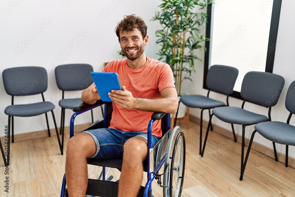 Young hispanic man using touchpad sitting on wheelchair at clinic waiting room. Young hispanic man using touchpad sitting on wheelchair at clinic waiting room.