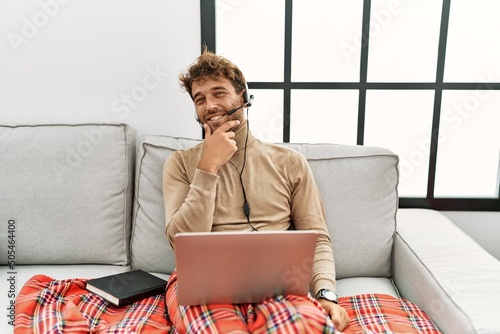 Young handsome man with beard wearing operator headset working from home looking confident at the camera smiling with crossed arms and hand raised on chin. thinking positive.