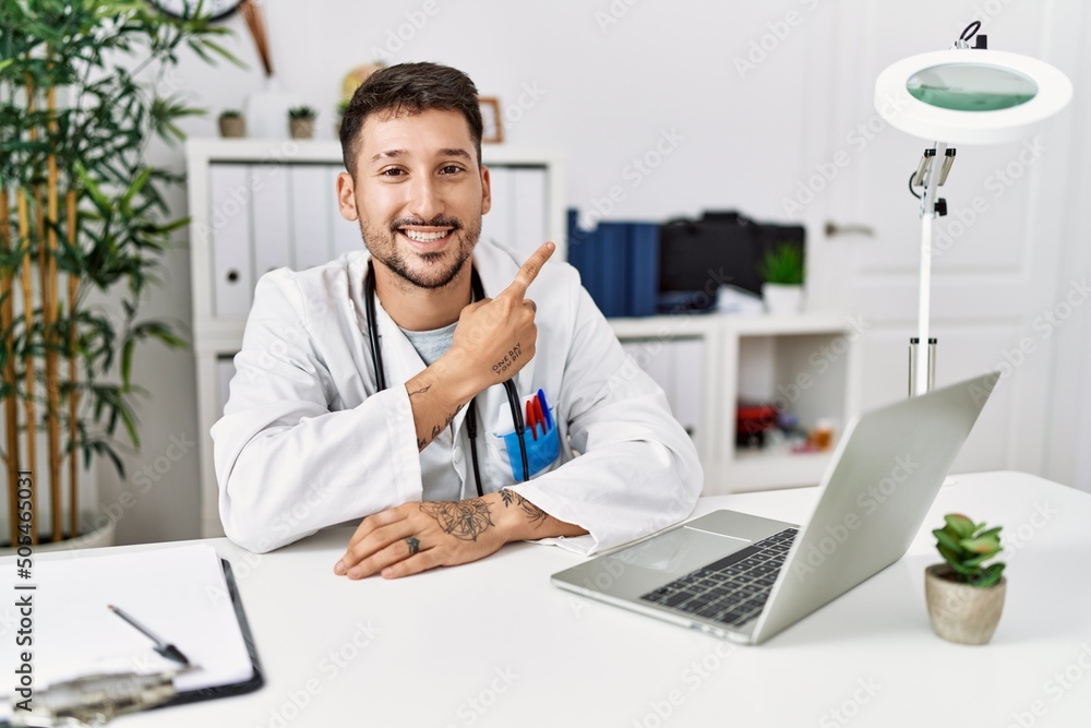 Young doctor working at the clinic using computer laptop cheerful with a smile of face pointing with hand and finger up to the side with happy and natural expression on face