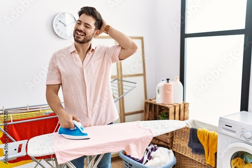 Young man with beard ironing clothes at home smiling confident touching hair with hand up gesture  posing attractive and fashionable