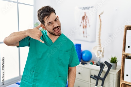 Young man with beard working at pain recovery clinic smiling doing phone gesture with hand and fingers like talking on the telephone. communicating concepts.