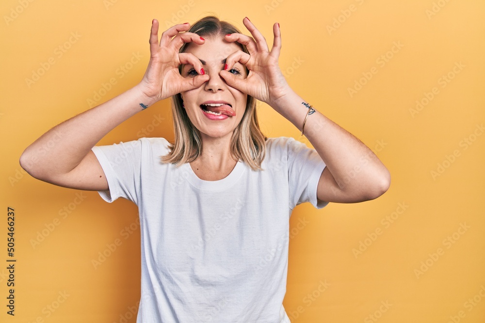 Beautiful caucasian woman wearing casual white t shirt doing ok gesture like binoculars sticking tongue out, eyes looking through fingers. crazy expression. Beautiful caucasian woman wearing casual white t shirt doing ok gesture like binoculars sticking tongue out, eyes looking through fingers. crazy expression.