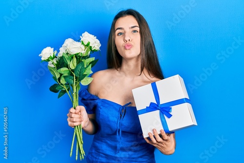 Young brunette teenager holding anniversary present and bouquet of flowers looking at the camera blowing a kiss being lovely and sexy. love expression.
