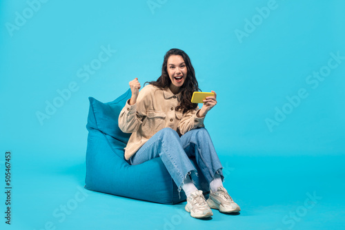 Glad young european brunette female with phone sits on bagchair and makes victory gesture photo