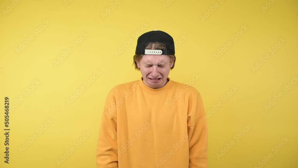 Sad guy in cap and sweatshirt with angry face isolated on yellow background, looks down and cries Sad guy in cap and sweatshirt with angry face isolated on yellow background, looks down and cries