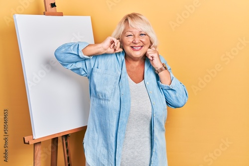 Middle age blonde woman standing by painter easel stand covering ears with fingers with annoyed expression for the noise of loud music. deaf concept.