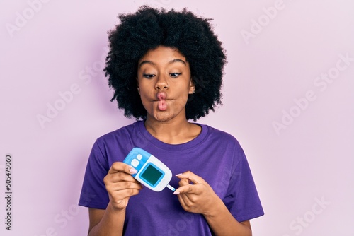 Young african american woman holding glucometer device making fish face with mouth and squinting eyes, crazy and comical.