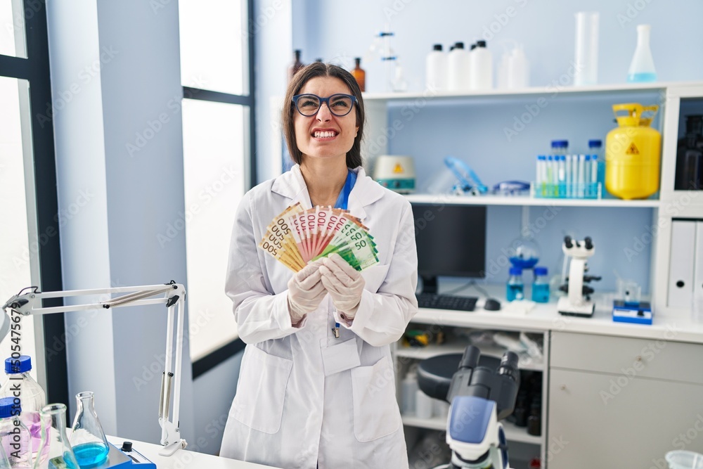 Young hispanic woman working at scientist laboratory holding money banknotes angry and mad screaming frustrated and furious, shouting with anger looking up. Young hispanic woman working at scientist laboratory holding money banknotes angry and mad screaming frustrated and furious, shouting with anger looking up.