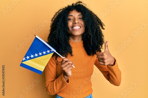 African american woman with afro hair holding bosnia herzegovina flag smiling happy and positive, thumb up doing excellent and approval sign photo