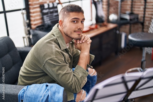 Young hispanic man musician smiling confident sitting on sofa at music studio