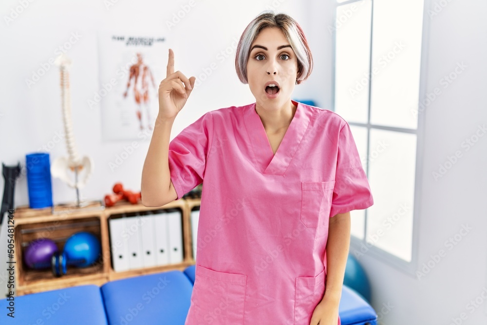 Young nurse woman working at pain recovery clinic pointing finger up with successful idea. exited and happy. number one. Young nurse woman working at pain recovery clinic pointing finger up with successful idea. exited and happy. number one.
