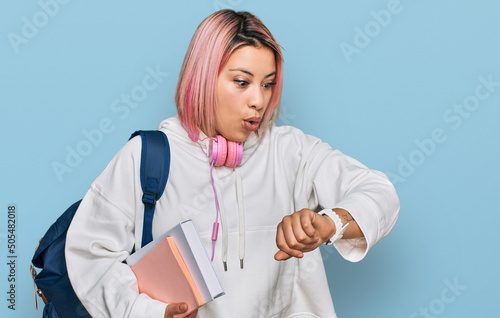 Hispanic woman with pink hair wearing student backpack and headphones looking at the watch time worried, afraid of getting late photo