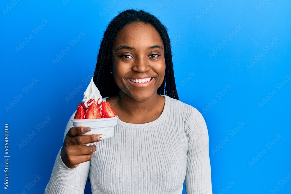 African american woman with braided hair eating strawberry ice cream looking positive and happy standing and smiling with a confident smile showing teeth African american woman with braided hair eating strawberry ice cream looking positive and happy standing and smiling with a confident smile showing teeth