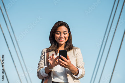 A happy elegant young latin woman stands and uses her phone to type an important message. A businesswoman using technologies outdoors.