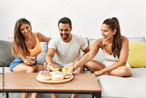 Three hispanic friends smiling happy eating doughnut at home.