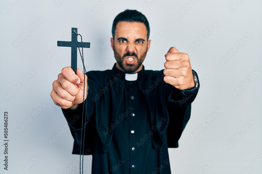 Handsome hispanic priest man with beard holding catholic cross annoyed and frustrated shouting with anger, yelling crazy with anger and hand raised Handsome hispanic priest man with beard holding catholic cross annoyed and frustrated shouting with anger, yelling crazy with anger and hand raised
