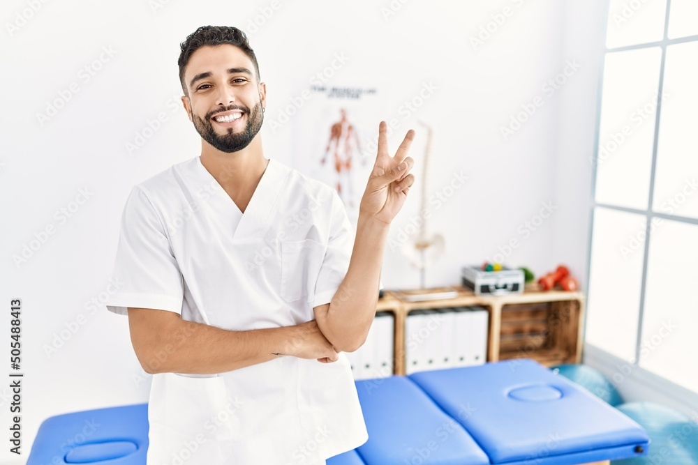 Young handsome man with beard working at pain recovery clinic smiling with happy face winking at the camera doing victory sign. number two.