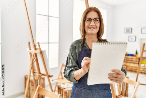 Middle age artist woman smiling happy painting on notebook at art studio.