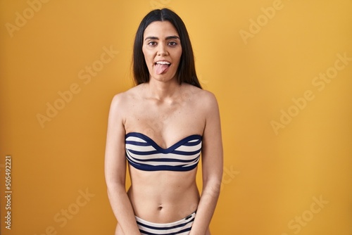 Young brunette woman wearing bikini over yellow background sticking tongue out happy with funny expression. emotion concept.