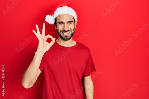 Young hispanic man wearing christmas hat smiling positive doing ok sign with hand and fingers. successful expression.