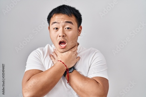 Young chinese man standing over white background shouting suffocate because painful strangle. health problem. asphyxiate and suicide concept.