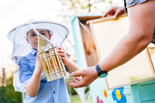 Selective focus of boy beekeeper in a protective suit holds a smoker in his hands. Father beekeeper passes his experience son. photo