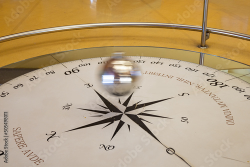 Foucault pendulum swinging on a compass rose. Pendulum blurred by movement. The text in latin 